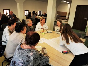 uczniowie i seniorzy prowadzą dyskusję w jednym ze stolików. Scena ma miejsce w sali budynku Centrum Aktywności Społecznej