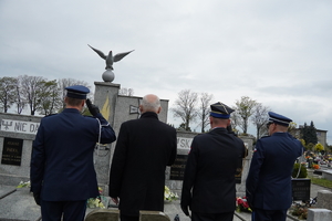 policjanci, strażak oraz starosta stoją przed pomnikiem na cmentarzu parafialnym, mundurowi salutują. Zdjęcie wykonane od tyłu