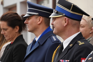 stojący obok siebie goście obchodów Dnia Strażaka, wśród nich policjant w mundurze galowym. Osoby stoją przed budynkiem domu kultury