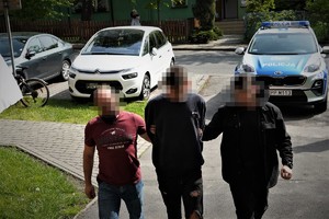 na zdjęciu widać trzech mężczyzn, którzy wchodzą do budynku komendy. Mężczyzna w środku to osoba zatrzymana. Za nimi na parkingu stoją samochody