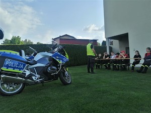 na pierwszym planie motocykl policyjny, dalej policjant i słuchacze szkolenia. Dzieci siedzą na ławce