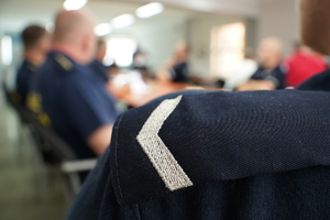 pagon na koszulce policjanta, który wraz z pozostałymi mundurowymi siedzi na sali odpraw w komendzie