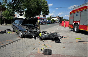 droga, leżący na jezdni rozbity motocykl i samochód koloru zielonego. Za nimi wóz straży pożarnej