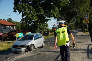 policjant drogówki prowadzi oględziny miejsca wypadku, za nim rozbity samochód