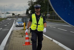 policjant drogówki niesie pachołek do zabezpieczenia trasy. Scena ma miejsce przy skrzyżowaniu dróg krajowych