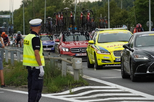 po lewej stoi policjant drogówki, po prawej peleton Tour de Pologne oraz samochody sponsorów