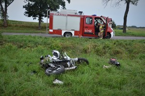 leżący na trawie rozbity motocykl, za nim na drodze stoi wóz straży pożarnej