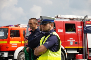 dwaj policjanci, jeden z nich ma założoną odblaskową kamizelkę, za nimi stoją wozy straży pożarnej