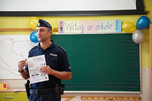 policjant w sali lekcyjnej, za nim zielona tablica z napisem WITAJ SZKOŁO