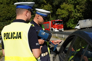 policjanci drogówki podczas kontroli samochodu osobowego, w tle widać wóz straży pożarnej