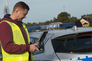 mężczyzna w odblaskowej kamizelce steruje dronem, stoi obok oznakowanego radiowozu